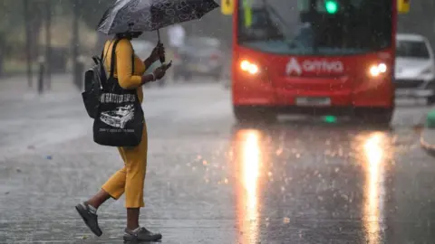 How to stay safe and what to do in a power cut Getty Images A woman wearing a yellow top and trousers and carrying an umbrella crosses the road in the pouring rain in front of a red double decker bus