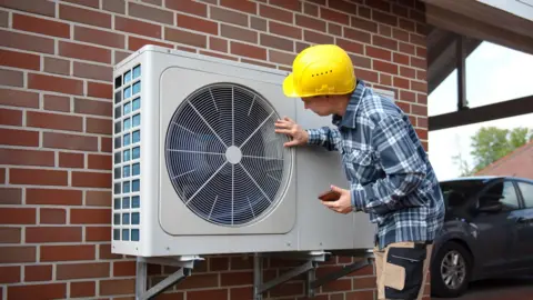 Heat pumps and EVs making progress, UK climate advisers say Getty Images Heat pump being installed on a red brick wall by a man in a yellow helmet and blue checked shirt.