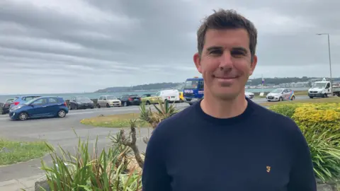 Family shocked at rental costs in Guernsey BBC Tom Pinsard is smiling at the camera as he stands on a garden. Behind him is a road which has cars parked along it next to a harbour wall with the sea extending beyond. He has short brown hair and is wearing a navy crew cut jumper.
