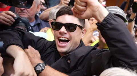 Rapper released on unconditional bail over terror charge PA Media Móglaí Bap (Naoise Ó Cairealláin) outside court, wearing a black jacket and sunglasses. He is surrounded by crowds of people.