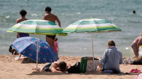 Health alerts come into force ahead of second heatwave PA Media People sunbathe on a beach beneath umbrellas because of the sun.