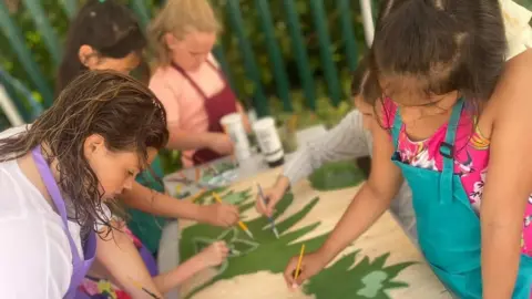 Bradford families urged to sign up to 'holiday hunger' clubs YACAD A group of young girls painting a large piece of artwork which is laid out on a table in front of them. It's a shared activity with them all working on the same piece. They hold small paintbrushes and wear colourful aprons.