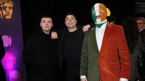 Rapper released on unconditional bail over terror charge Getty Images Liam Óg Ó hAnnaidh, Naoise Ó Cairealláin and J.J. Ó Dochartaigh aka Mo Chara, Móglaí Bap and DJ Próvaí of Kneecap at the BAFTAS. Liam has short dark hair wearing black clothing. Naoise has dark wavy hair with his arms leant on the shoulders of Liam and JJ, he is wearing black clothing and rings. JJ is wearing a tri colour balaclava and tri colour suit. They are standing next to a BAFTAS sign.