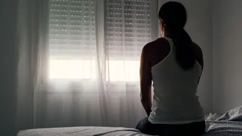 National Crime Agency to launch operation targeting grooming gangs Getty Images A woman has her back to the camera and is sitting on the edge of a bed in a dark room, facing the window. She has her hair tied back into a ponytail and is wearing jeans and a white tank top.