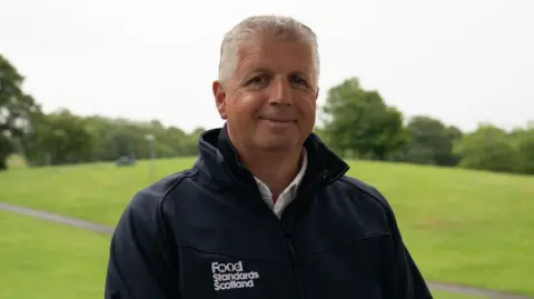 Fraudster jailed for selling fake 'Scottish-grown tea' A man with white hair wearing a black jacket with "Food Standards Scotland" emblazoned on it smiles at the camera. He is standing in an open green space like a park