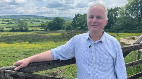 Warning over 'dirty secret' of toxic chemicals on farmers fields BBC/Jonah Fisher Farmer Richard Smallwood is wearing a blue and white checked shirt and leaning on a fence in front of lush green countryside.