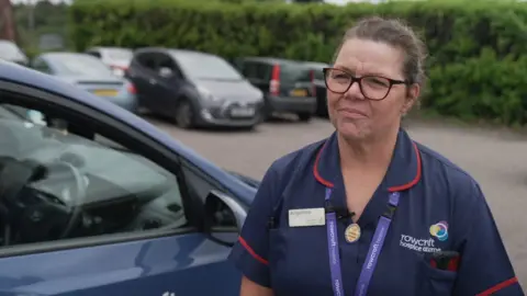 Hospice staff tell BBC what they think about bill Angelina Blair, who is wearing a dark blue nurses uniform, has brown hair tied back and is wearing brown-rimmed glasses.