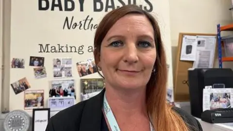 Wollaston charity supports one in 16 babies across the county Annabel Amos/BBC Sabrina Oakey with long light brown hair flowing down her left shoulder. She is smiling at the camera and wearing a black top and white lanyard. There are photos on the wall behind her and parts of the words "Northampton" and "making" are visible.
