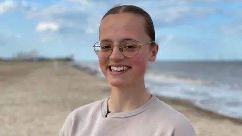 New drug offers potential cure for ultra rare inherited condition BBC Mary Catchpole, smiling with her hair tied back and wearing glasses. She is standing on a beach near her home in Norfolk