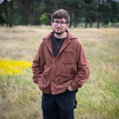 A preacher asked me out and then started stalking me BBC A man in a brown jacket with black trousers stands with his hands in his pockets with a long green meadow and trees in the background.