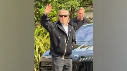 Bruce Springsteen and Paul McCartney in surprise Liverpool duet Anderson Tombini Paul McCartney wearing black sunglasses, a black jacket, white shirt, and jeans, waves as he stands in front of a black BMW car.