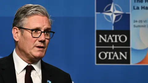 PM defends leadership amid growing welfare rebellion PA Media Sir Keir Starmer stands speaking at a Nato summit, wearing a dark suit, white shirt, and dark tie. Behind him is a blue backdrop featuring the Nato logo