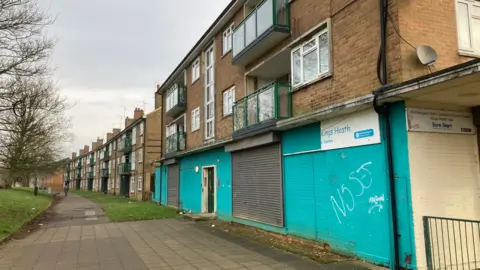 Cost of living roadshows to tour West Northamptonshire Martin Heath/BBC Street of terraced three-story flats, curving round to the left. There is a grassed area to the left with trees and a wide pavement separating it from the buildings.
There is a boarded-up council building to the right - a sign is visible which says King's Heath Centre and has a Northamptonshire County Council logo.