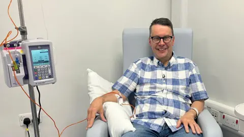World-first 'Trojan horse' therapy to be given on NHS Paul wearing a blue and white checked shirt is sat in a chair, bare arm is resting on a cushion while an orange tube connects a cannula to a pump.