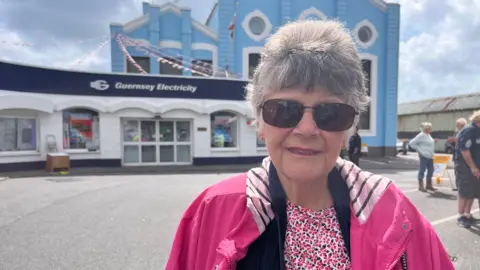 'We sit in the dark to save money on electricity in Guernsey' BBC A woman with short grey hair, wearing sunglasses and a pink patterned T-shirt and pink coat stands in front of a blue and white Guernsey Electricity building.