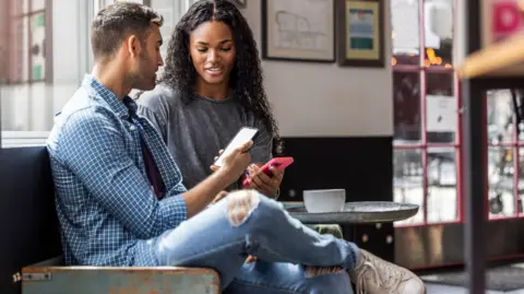 Dating app scam victims told 'don't suffer in silence' Getty Images Man and a woman sit in a coffee shop each looking at a smartphone.