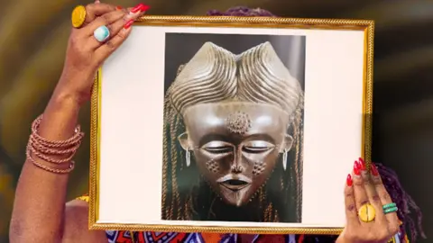 Women’s History Museum Zambia Samba Yonga from Women's History Museum of Zambia holds up a frame over her face showing a photograph of a sacred mask with Sona symbols etched on to its surface, each telling stories of women's significance, wisdom, and the vital knowledge they carried.