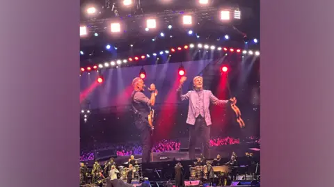 Bruce Springsteen and Paul McCartney in surprise Liverpool duet RossEFC95 / X A big screen and stage, showing Sir Paul McCartney holding his hands up to the crowd and holding a guitar. Bruce Springsteen is standing to his left clapping, holding a guitar and smiling. There is red and blue lighting on stage with other musicians.