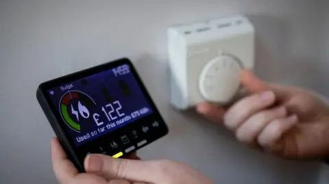Why are electricity prices going up in Guernsey? EPA A pair of hands - one hand is holding a smart meter showing the money spent on electricity and another is adjusting a thermostat.