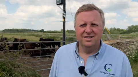 Glastonbury farmer 'makes more money from weddings than farming' A man in a light blue polo shirt with blonde hair is standing in front of the gate to a field where a herd of black cows is grazing.
