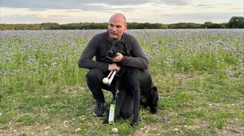 How to keep your dog safe in a heatwave BBC BBC correspondent Sean Dilley pictured in a field on a bright day, crouched down with his arms around his black guide dog Shawn
