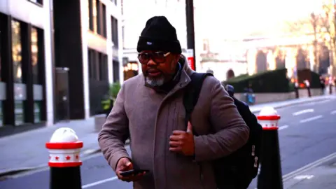 NHS trust and medic guilty of failings Benjamin Aninakwa pictured walking towards the camera on his way along a busy road into the Old Bailey, wearing glasses, a hat and coat, and carrying a shoulder bag.