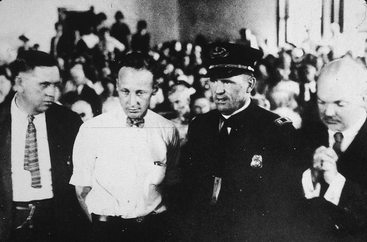Pop culture helped fan the flames of the Scopes ‘monkey trial’ 100 years ago − and ever since A black and white photograph of four men standing amid a crowded room, including one younger man in a white, short-sleeve shirt and bow tie.