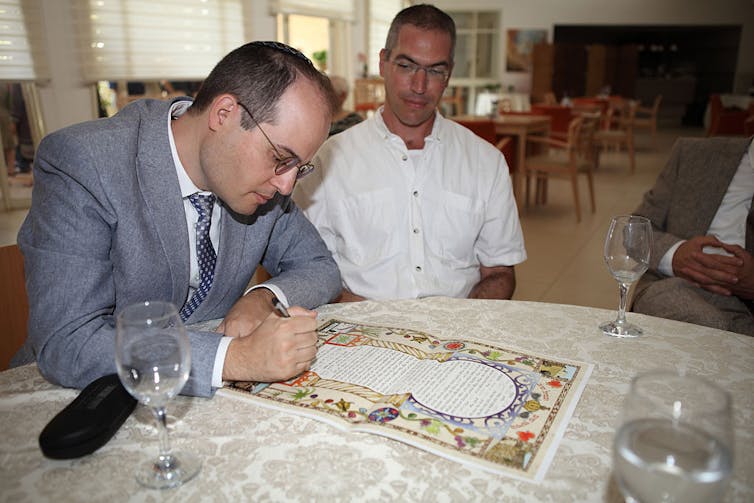 From the marriage contract to breaking the glass under the chuppah, many Jewish couples adapt their weddings to celebrate gender equality A man in a blue-gray suit signs a colorfully decorated piece of paper as another man in a white shirt watches.