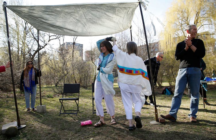 From the marriage contract to breaking the glass under the chuppah, many Jewish couples adapt their weddings to celebrate gender equality Two women wearing white clothes and prayer shawls dance under a simple canopy in a park as a few people look on.