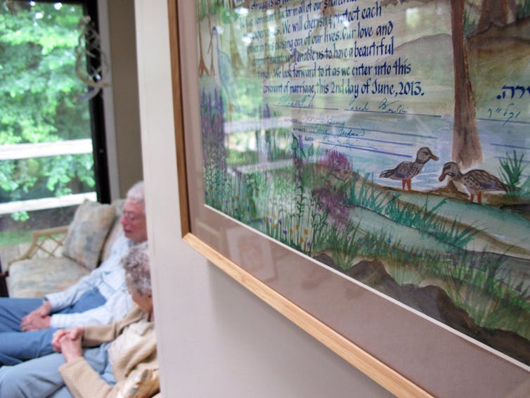 From the marriage contract to breaking the glass under the chuppah, many Jewish couples adapt their weddings to celebrate gender equality A colorful, framed drawing on a white wall, with two older women barely visible sitting on a couch at the back of the room.