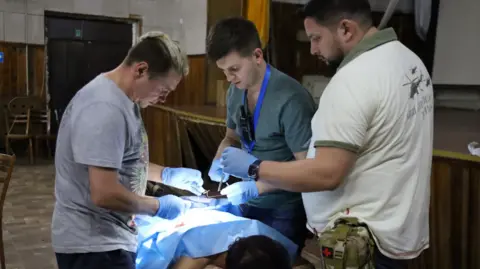 The doctor fighting for women's health on Ukraine's front line Serhii Baksheiev A woman lies face down on a table with a blue operating sheet over her body with three medics wearing gloves standing around her.