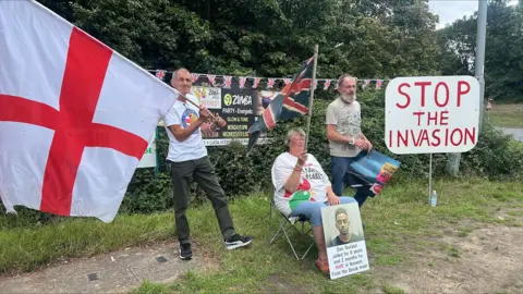Hundreds of protesters gather at asylum hotel in Norwich area Richard Knights/BBC Picture shows three men, one holding a St George Flag and the other is sitting down while holding a poster. The third man is standing next to a sign that reads 'Stop the invasion'.