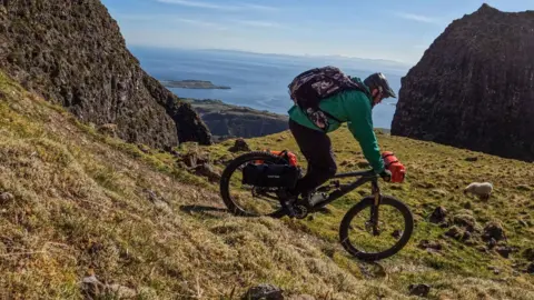 Mountain biker completes epic bid to ride down all 282 Munros Grizzly Munro Diaries A person on a mountain bike rides down a grassy hill overlooking a body of water.