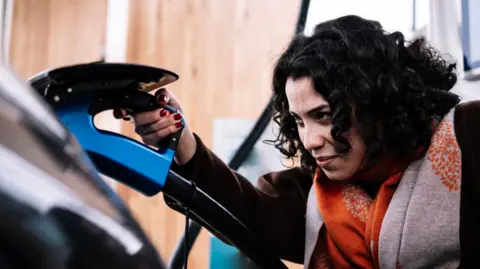 Electric vehicles to be discounted in government scheme Getty Images Woman with dark hair plugs in electric vehicle to charging point