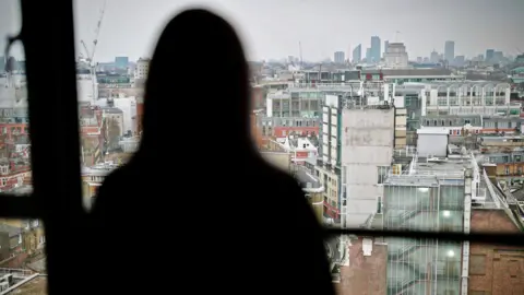 How MI5 piled falsehood on falsehood in court in the case of a spy who abused women Beth, pictured in a blurred silhouette against a high window, looking out onto tall buildings stretching into the distance on an overcast day