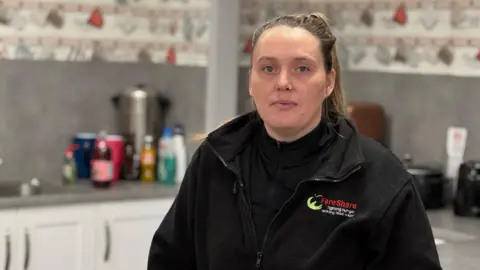 'Food demand in Cumbria is unprecedented' BBC Naomi Winter with brown hair tied back in a ponytail, with a nose ring, wearing a black fleece that has the words 'fareshare' on the chest, stands in a kitchen.