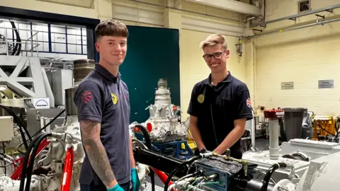 Defence firms 'confident' hundreds more jobs will come BBC Two men in a factory smile at camera. There are various engine parts behind them. Both are wearing blue tops, and are younger workers. The factory walls are painted yellow.