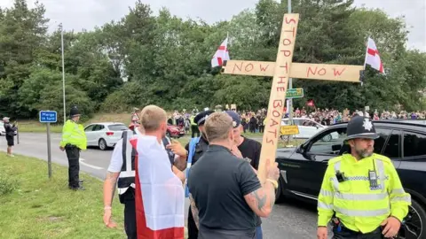 Hundreds of protesters gather at asylum hotel in Norwich area Alex Dunlop/BBC A man holding a large wooden cross that reads 'stop the boats now' with two St George flags attached on either sides of them. There are police officers also at the scene