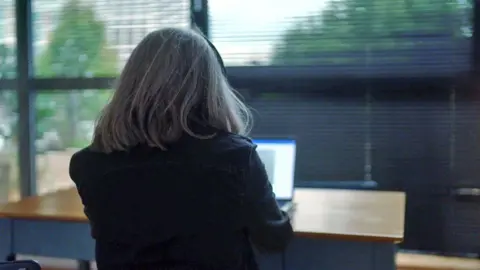 Dubious sales tactics at two leading estate agencies uncovered by BBC investigation Picture of the Purple Bricks whistleblower taken from behind. She is sitting a a wooden desk with a laptop, in front of a large window which has metal blinds. She has shoulder-length straight grey hair.