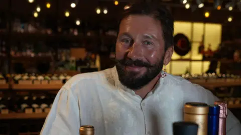Why small-time criminals burned a London warehouse for Russia's mercenary group Wagner A man with a beard and moustache smiles at the camera. He is in a wine shop with bottles behind him on the shelf