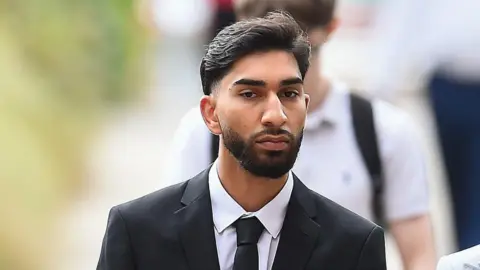 Man guilty of Manchester Airport attack on police officers PA Media A young man with a dark beard wears a white shirt and a black tie and jacket and walks along a street.
