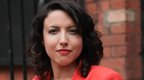 'Why I'm one of millions of UK adults not paying in' Victoria Olsena Victoria looking straight at the camera in business attire of a bright peach blazer, with dark shoulder length hair, wearing red lipstick and a gold chain.