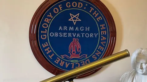 230 years of unbroken weather recordings A large Armagh Observatory crest with a wooden frame on a wall. A shiny brass telescope on a shelf under the crest, along with a with bust of a Georgian era man.