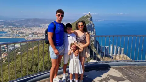Package holidays to Spain, Cyprus and Turkey soar in price Francesca Ramsden A family of four stand on a platform overlooking a mountain and coastline. On the left is a tall man wearing a blue top and sunglasses, in the middle is a boy with a black cap and white top, on the right is a woman in sunglasses and an animal print top and in front is a young girl in a white dress
