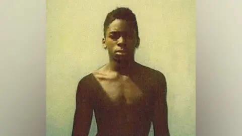 Broward State Attorney’s Office A police image of Mark Gordon taken at the time of his arrest in 1989. He is standing bare-chested against a yellow background and is looking directly ahead. 
