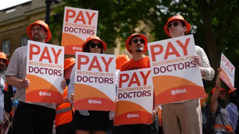 I deeply regret the position we're in, Streeting tells resident doctors PA Media Three young men and a woman stand in a line at a doctors' strike. They are all wearing identical orange bucket hats and holding placards towards the camera which say 'pay restoration for doctors'. A tree and a crowd of protesters are visible behind them.