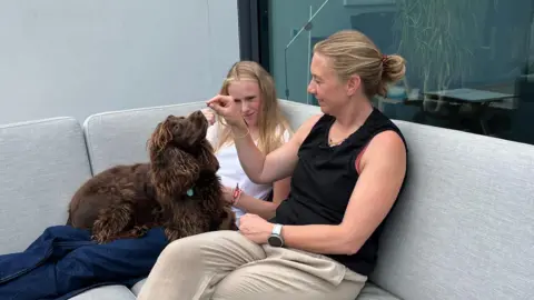 Babies from three people's DNA prevents heriditary disease BBC/Josh Elgin Kat Kitto, sitting on a grey corner sofa wearing a black vest top, with her daughter Lily who is wearing white. Kat is feeding Monty, a ginger coloured long-haired dog, who is sitting on Lily's legs