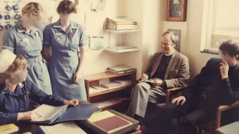 Police investigate sex abuse claims against men linked to Church D J Photography Three nurses in blue uniform crowd around a desk in an old office, with the closest woman sat pointing at a page of a folder. Over the other side a younger Anthony Pierce is seen sitting with his leg crossed; he is wearing grey trousers, a brown jacket and has a dog collar on. He looks relaxed and smiles as he takes notes. To his left is a man wearing black who is sat leaning and listening.