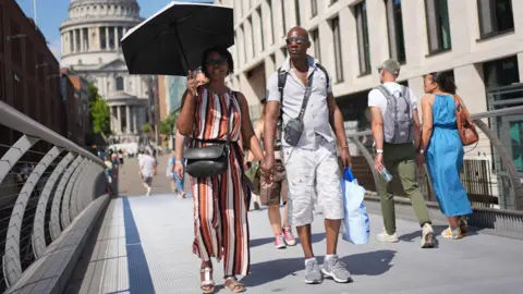 PA Media A woman wearing a black, white, yellow, and orange striped jumpsuit holds an umbrella in the sun. A man wearing a grey shirt and white shorts holds her hand and walks with her.