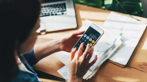 What is an Isa and how might the rules change? Getty Images Woman sits at a desk with paperwork and a laptop in front of her. A smartphone in her hand has a calculator on the screen.